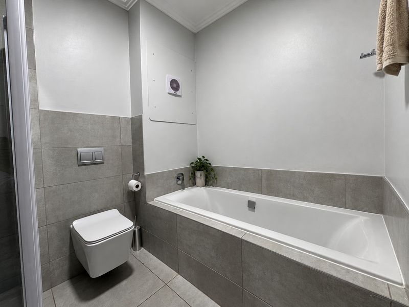 A modern, clean bathroom featuring a built-in bathtub, wall-mounted toilet, and neutral grey tiling throughout.