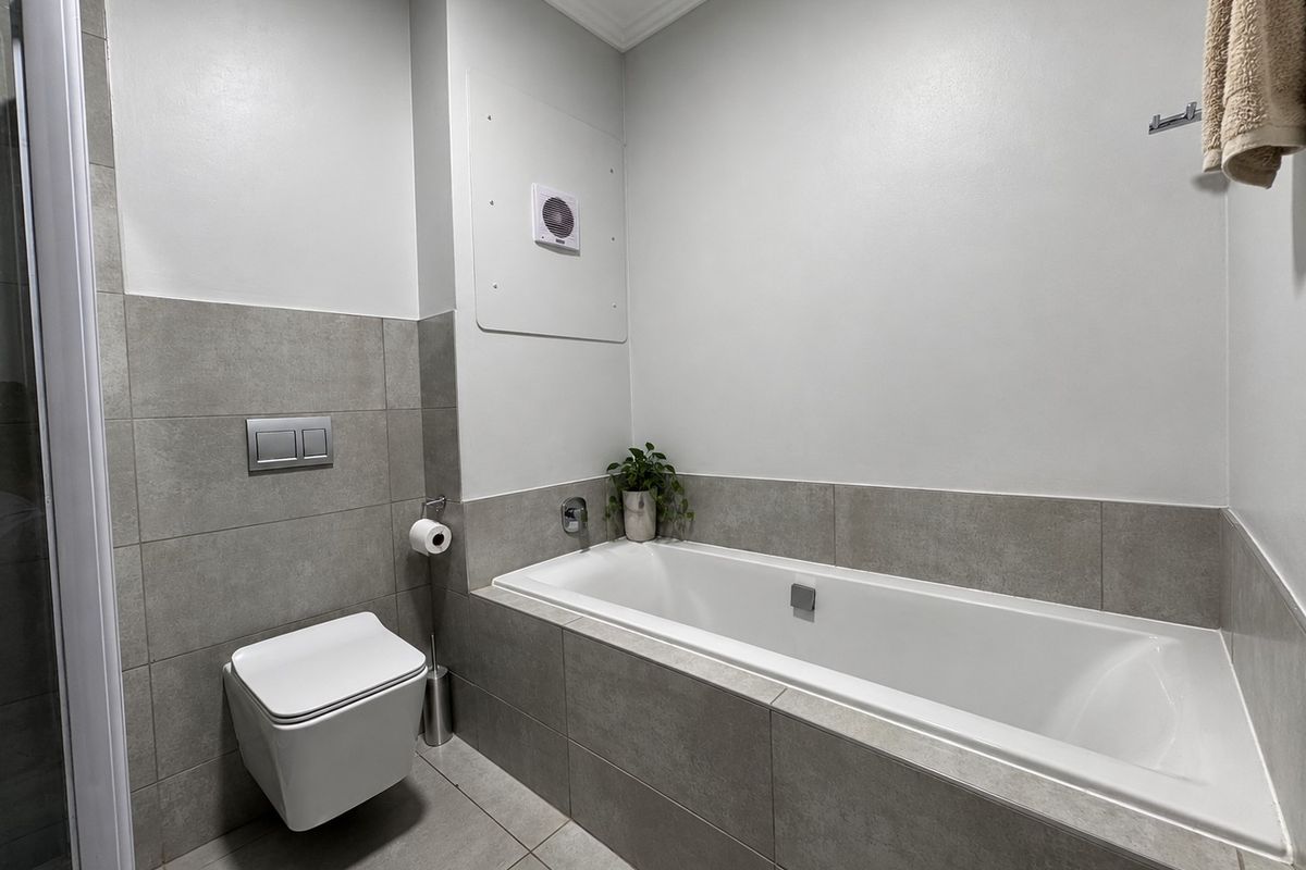 A modern, clean bathroom featuring a built-in bathtub, wall-mounted toilet, and neutral grey tiling throughout.