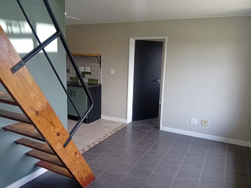 Open plan living area featuring a wooden staircase, tiled flooring, and a compact kitchen corner with cabinetry and tiled backsplash.