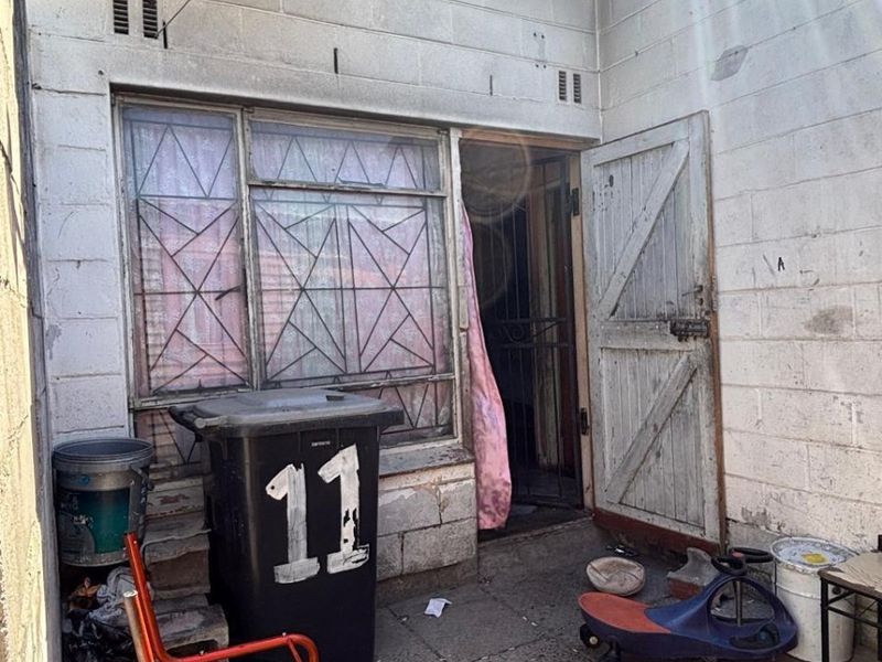 Rear exterior view of an apartment unit featuring concrete block walls, a metal security gate, and window burglar bars in need of maintenance.