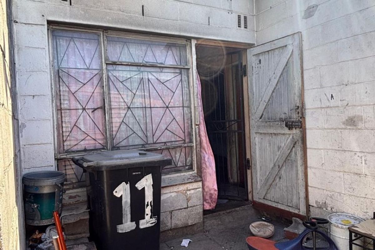 Rear exterior view of an apartment unit featuring concrete block walls, a metal security gate, and window burglar bars in need of maintenance.