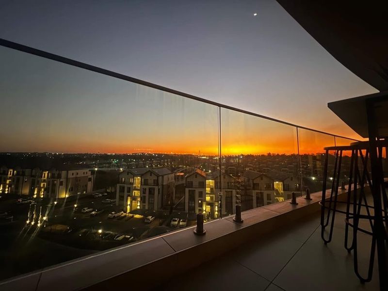 A modern penthouse balcony featuring a sleek glass balustrade and tiled flooring, offering an expansive sunset view over the surrounding residential estate.