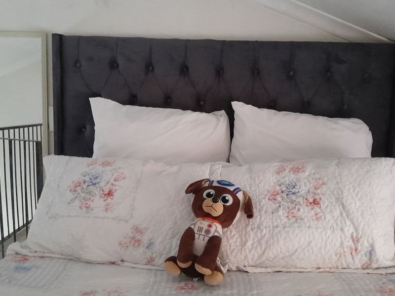 A cozy bedroom area featuring a dark tufted headboard and a sloped ceiling, overlooking a mezzanine level with a metal balustrade.
