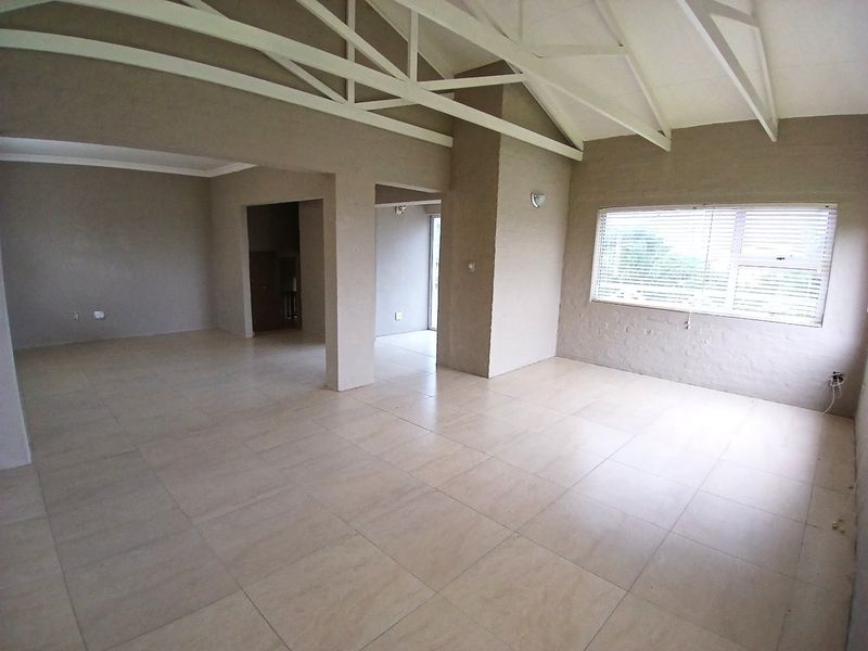 Spacious open-plan living area featuring vaulted ceilings with exposed white trusses, neutral tiled flooring, and large windows with fitted venetian blinds.