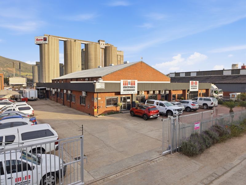 An industrial property featuring a brick facade, metal roofing, and a paved driveway, situated near large industrial silos with secure perimeter fencing.