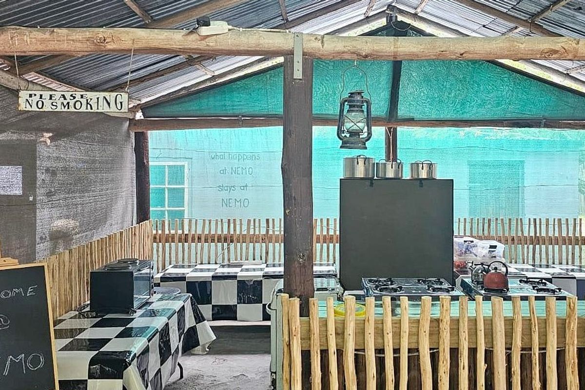 A rustic, open-air kitchen area featuring a gas hob, timber pole construction, and corrugated metal roofing, suitable for lodge-style outdoor catering.