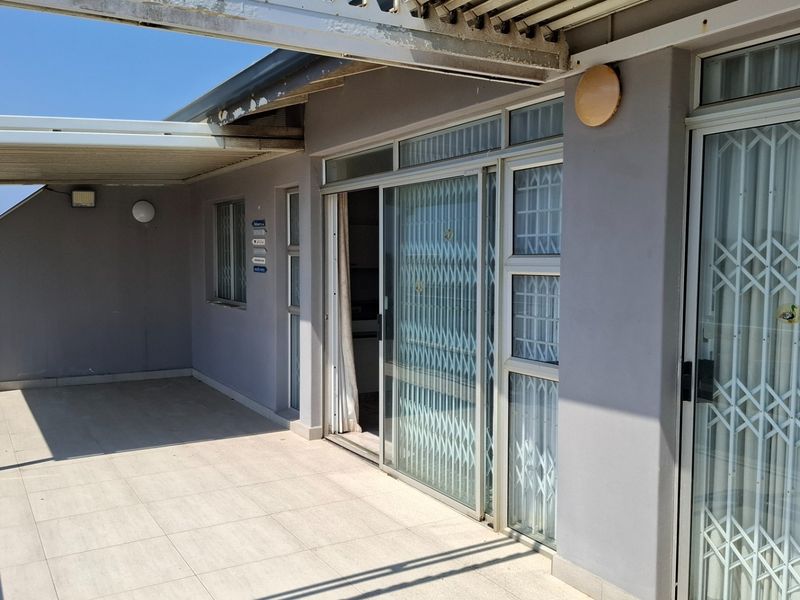 A spacious covered balcony featuring tiled flooring, sliding glass doors with security gates, and exterior wall lighting, offering a functional outdoor living space.