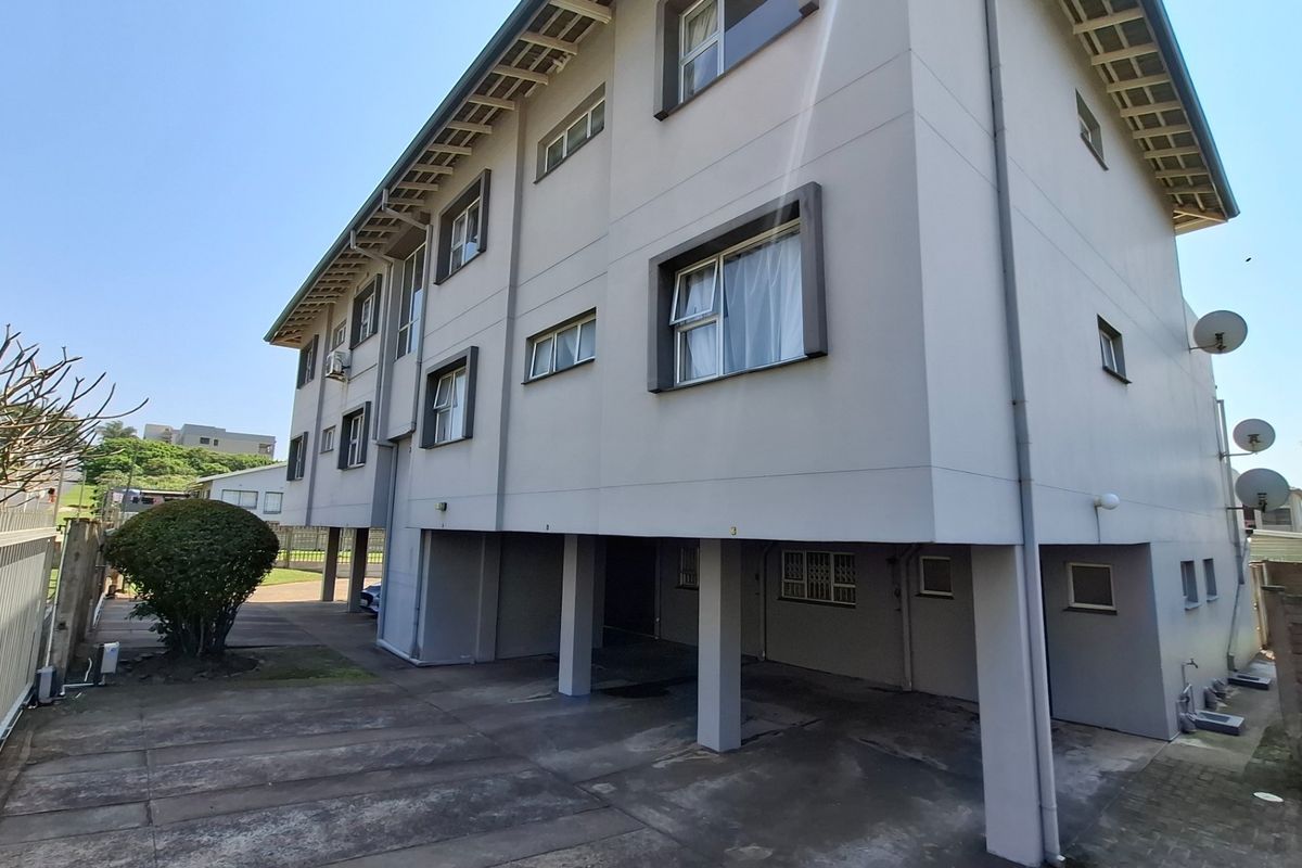 Rear view of a multi-story apartment building featuring a stilted ground level, concrete driveway, and exterior security fencing.
