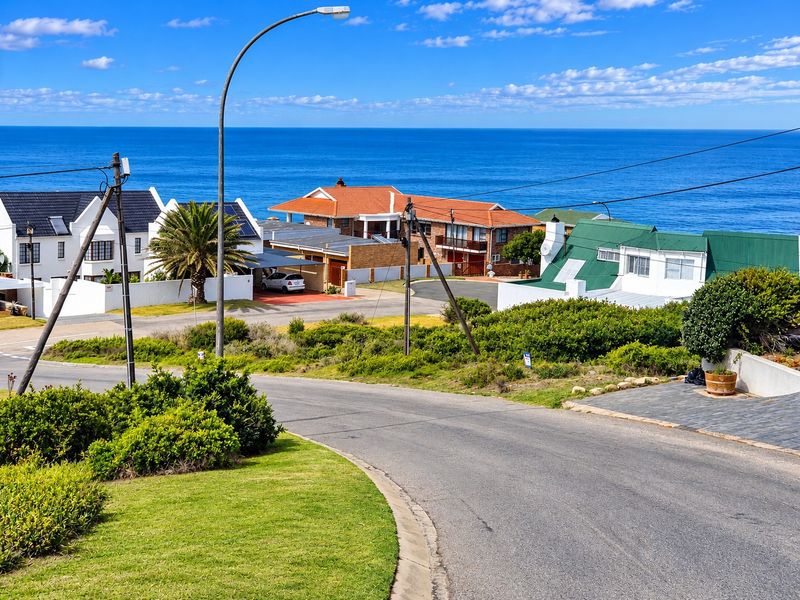 A street-level view of a residential neighborhood featuring paved roads, landscaped verges, and coastal views in the background.