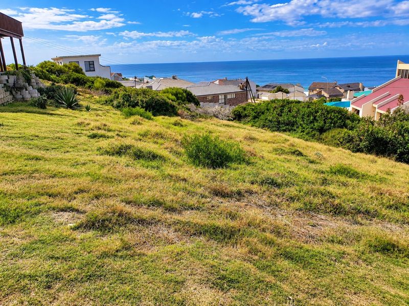 A scenic view of the ocean and surrounding residential neighborhood from a sloped, grass-covered vacant residential plot.