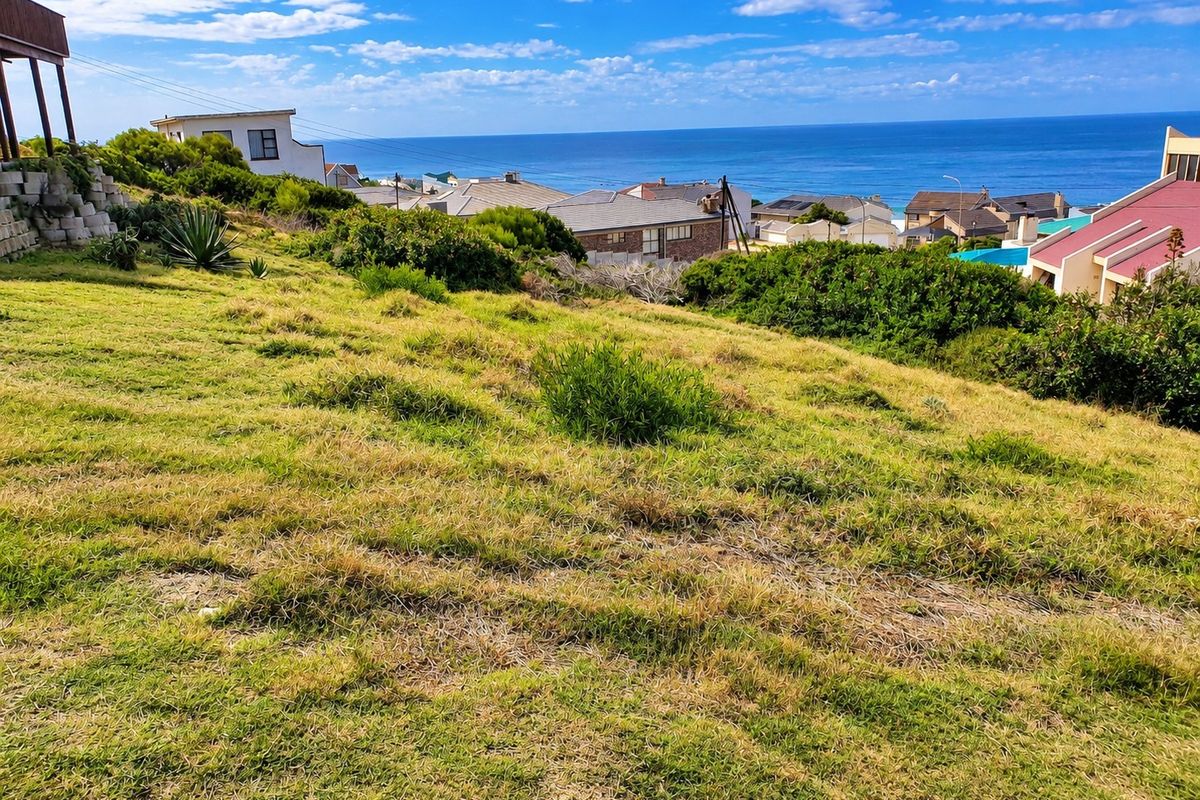 A scenic view of the ocean and surrounding residential neighborhood from a sloped, grass-covered vacant residential plot.