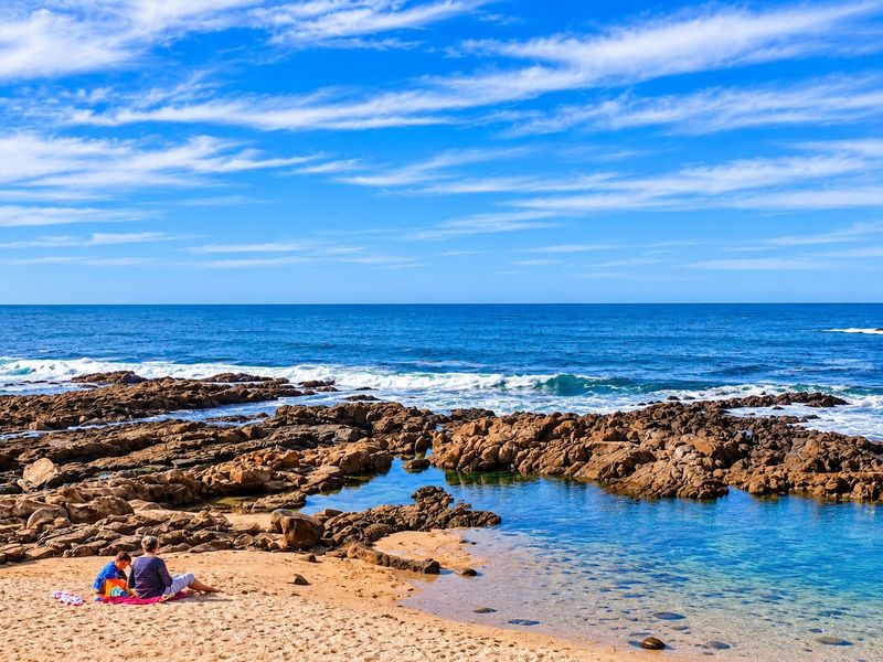 A scenic coastal view featuring rocky shoreline, clear blue ocean waters, and a sandy beach area.