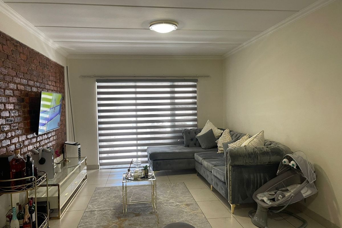 A comfortable living room featuring an exposed brick accent wall, neutral tiled flooring, and modern roller blinds, creating a warm and inviting atmosphere.
