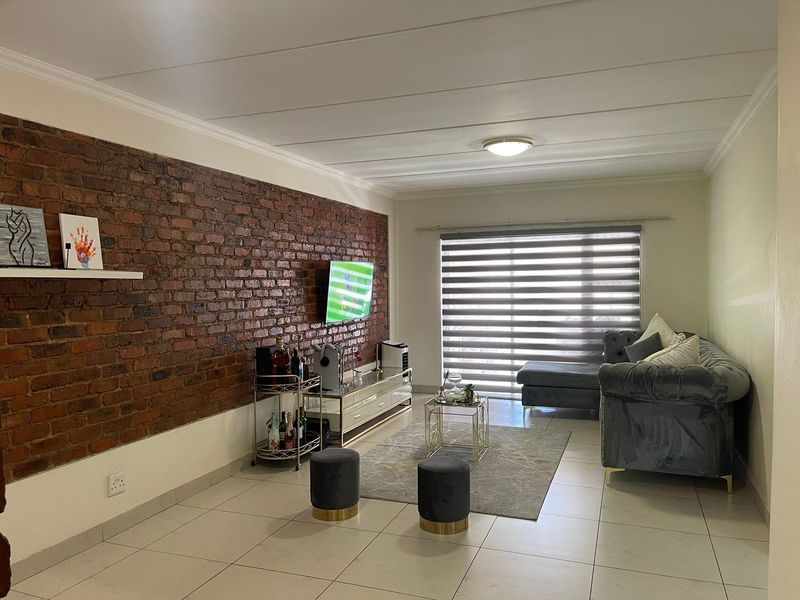 A well-maintained living room featuring a striking exposed brick feature wall, neutral tiled flooring, and modern roller blinds for adjustable natural light.