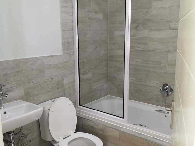 Modern bathroom featuring a wall-mounted basin, toilet, and a bathtub with a glass shower screen, finished with neutral wood-look wall and floor tiles.
