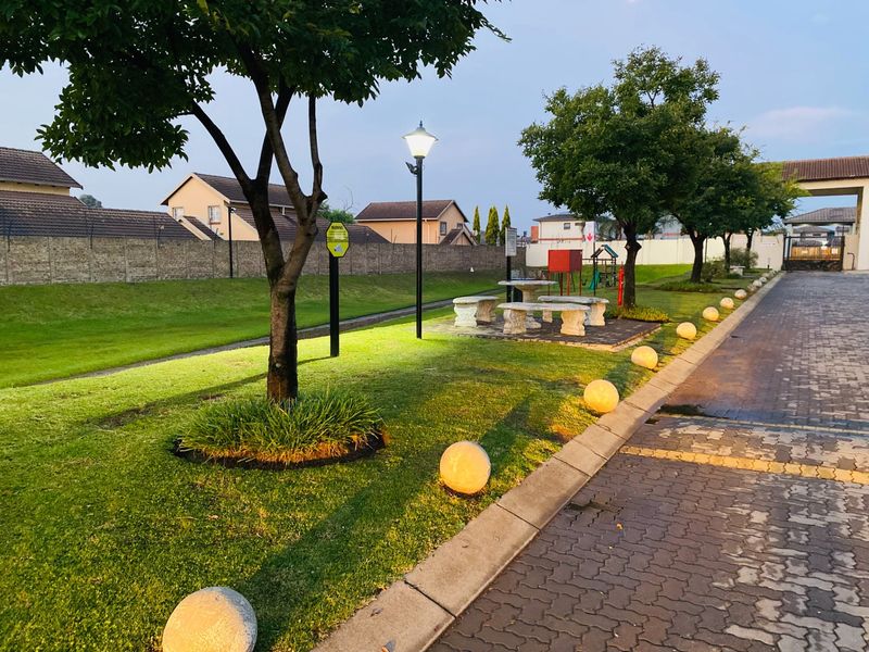 A well-maintained communal garden area featuring a paved driveway, manicured lawn, mature trees, and decorative garden lighting within a secure residential estate.