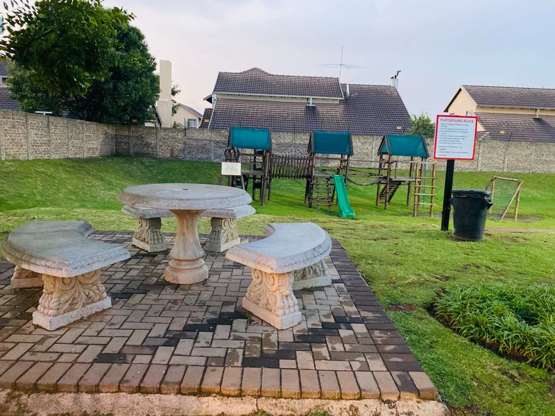 Communal garden area featuring a paved patio with stone seating, a lawn, and playground equipment enclosed by a perimeter wall.