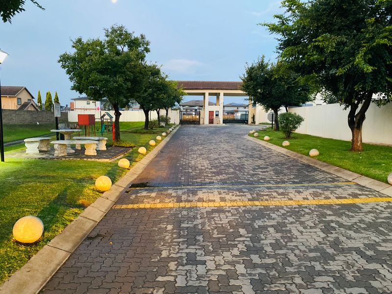 A well-maintained entrance to the townhouse complex featuring a paved driveway, secure gated access, and landscaped garden areas with established trees.