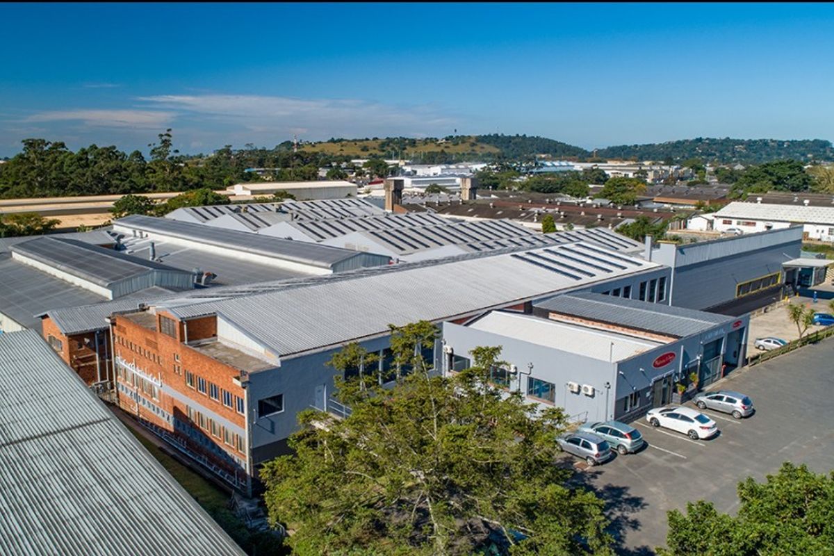 Aerial view of a large commercial industrial facility featuring extensive corrugated metal roofing, brick and painted masonry facades, and an adjacent asphalt parking area.