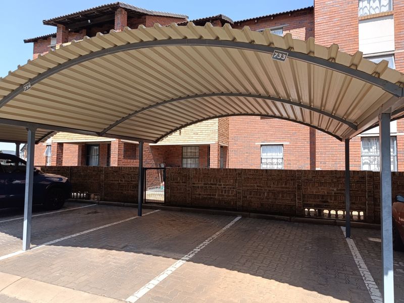 A covered parking carport with a metal roof structure and paved parking bays located within an apartment complex.