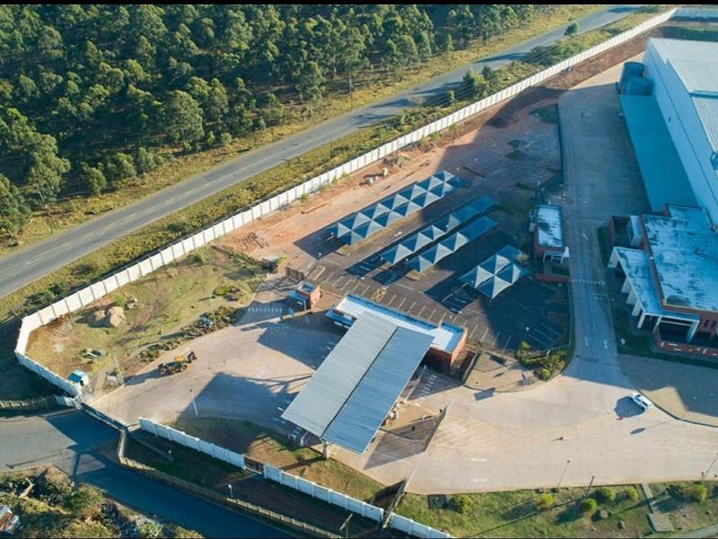 Aerial view of a commercial property featuring a large warehouse, paved parking area with carports, security gatehouse, and perimeter walling.