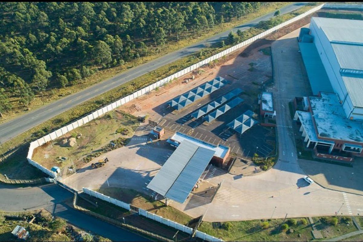 Aerial view of a commercial property featuring a large warehouse, paved parking area with carports, security gatehouse, and perimeter walling.