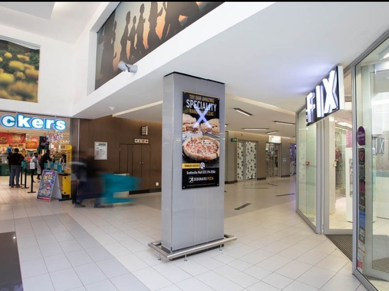 A well-lit retail shopping mall corridor featuring tiled floors, glass storefronts, and a central structural column with integrated advertising signage.