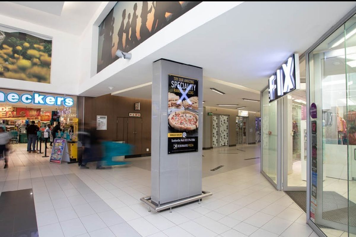 A well-lit retail shopping mall corridor featuring tiled floors, glass storefronts, and a central structural column with integrated advertising signage.