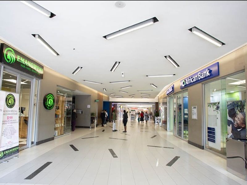 A well-lit, modern retail shopping centre corridor featuring tiled flooring, recessed ceiling lights, and multiple glass-fronted commercial storefronts.
