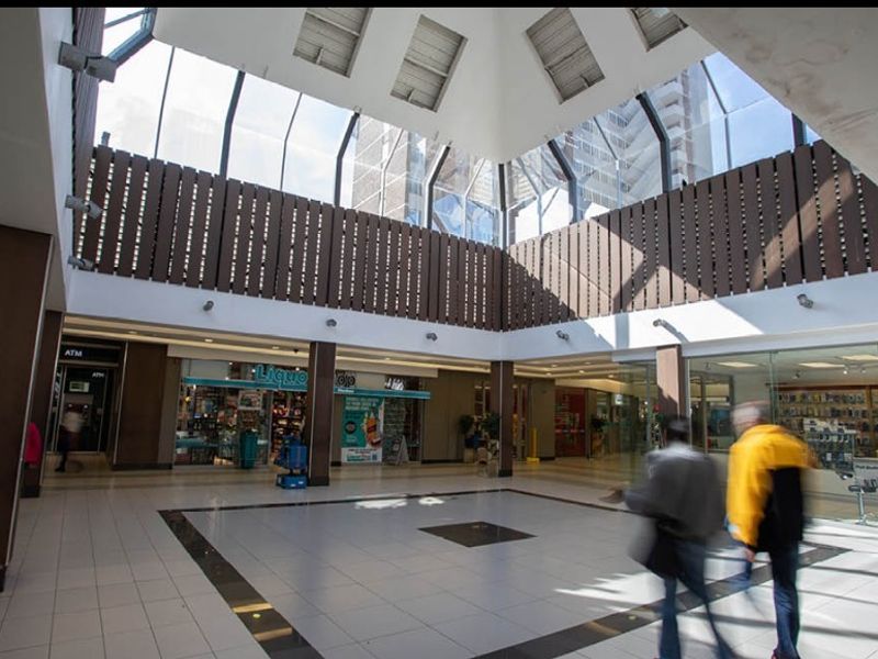 A bright, open-plan retail atrium featuring a large central skylight, tiled flooring, and multiple glass-fronted commercial units.