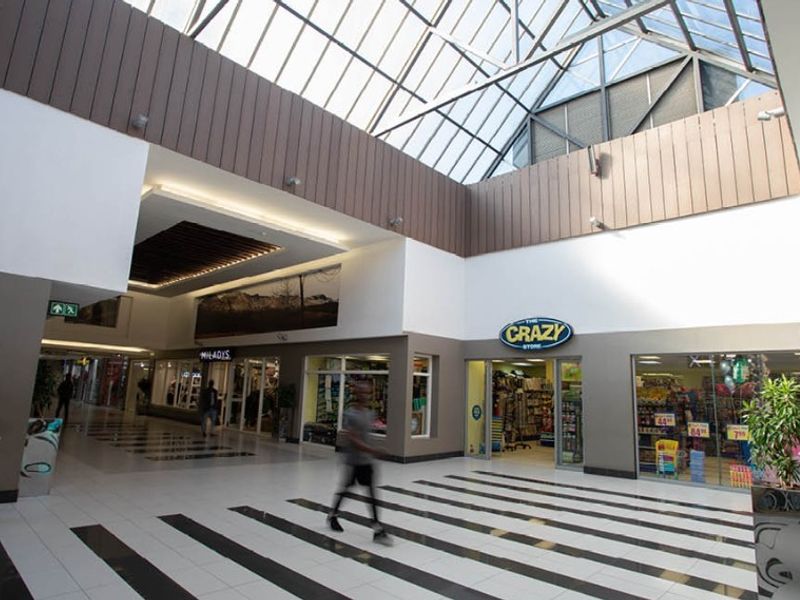 Interior view of a modern retail shopping center corridor featuring a large glass skylight, tiled flooring, and multiple storefronts.