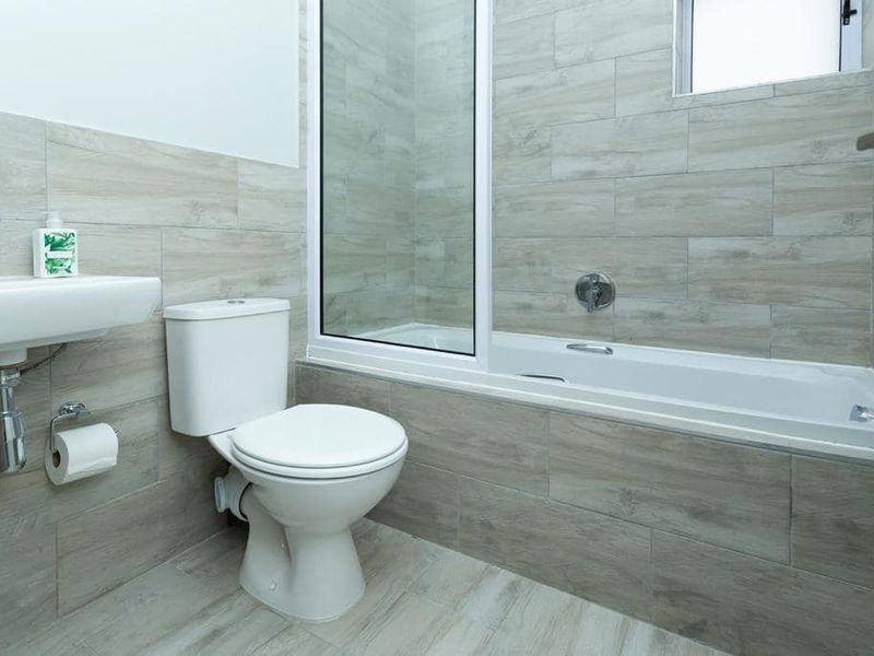 A modern, clean bathroom featuring neutral wood-look wall and floor tiles, a white bathtub with a glass screen, a toilet, and a wall-mounted basin.