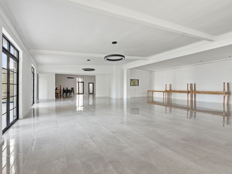 A spacious, bright open-plan office area featuring polished porcelain flooring, modern pendant lighting, and large black-framed glass doors providing ample natural light.