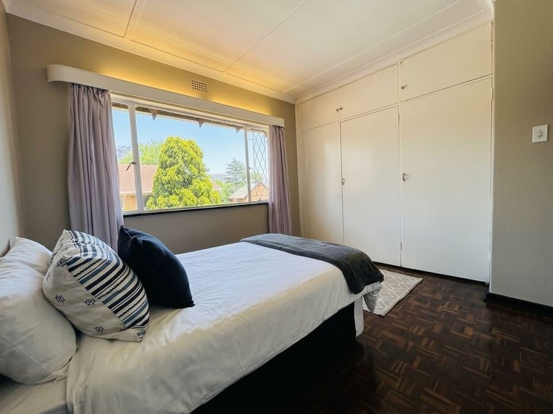 A well-maintained bedroom featuring classic parquet flooring, ample built-in cupboard storage, and a large window providing natural light and pleasant exterior views.