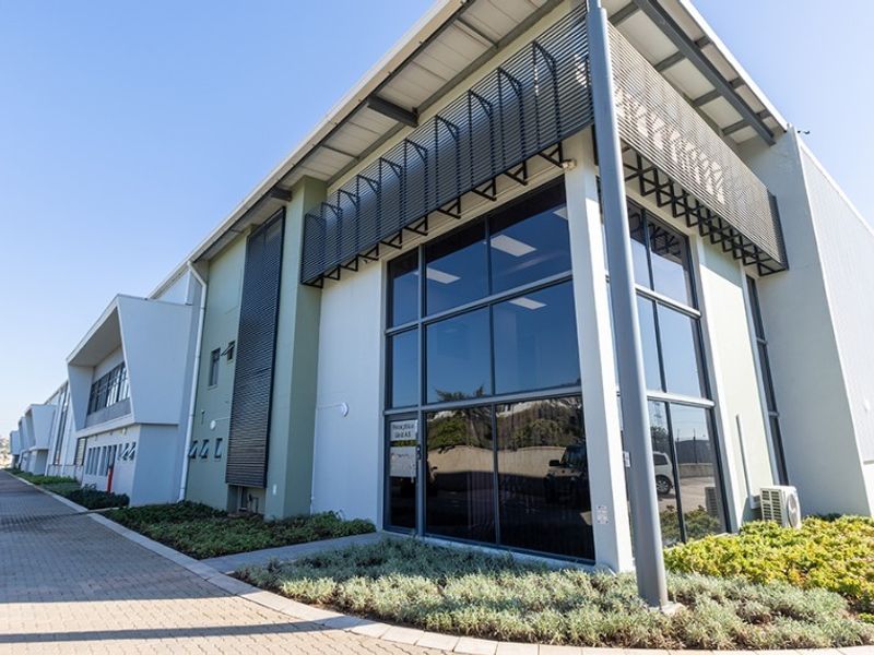 Modern commercial building exterior featuring a prominent glass facade, metal sunshades, and well-maintained paved driveway with landscaped garden beds.