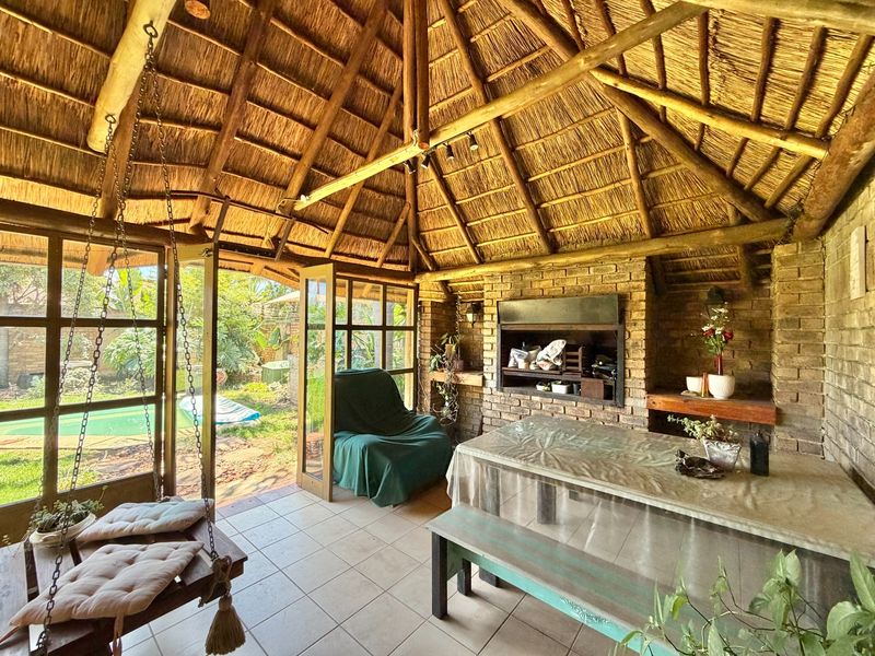Covered outdoor patio featuring a traditional thatched roof, exposed timber beams, built-in brick braai, and direct access to the garden and pool area.