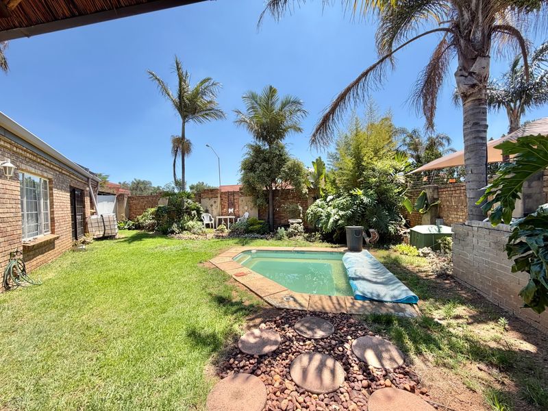 A private backyard featuring a swimming pool, established garden with palm trees, and brick boundary walls, offering a relaxed outdoor living space.