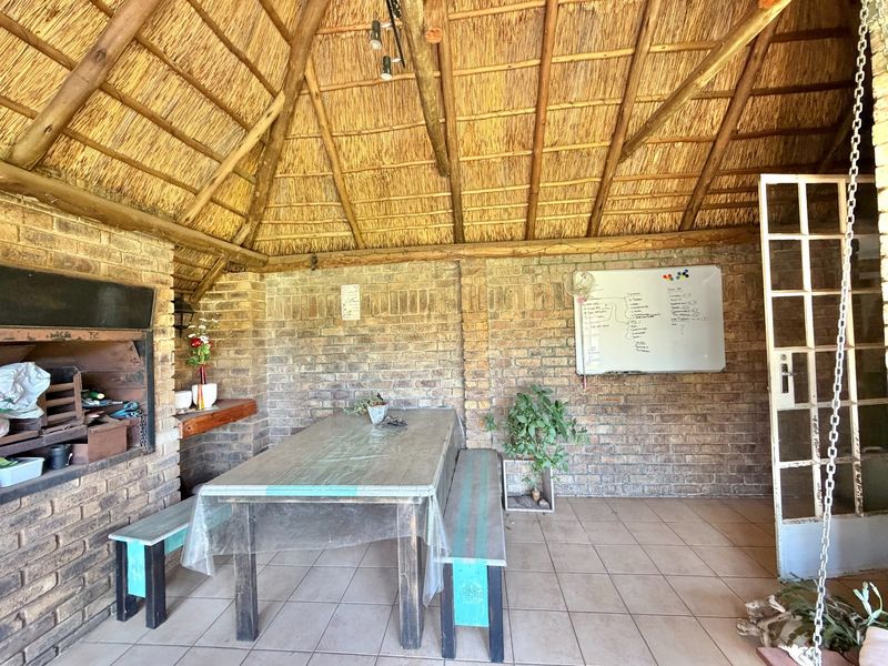 A covered outdoor patio area featuring a traditional thatched roof, exposed brick walls, and a built-in brick braai, perfect for outdoor entertaining.