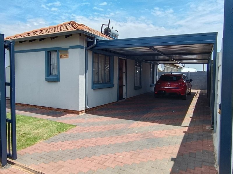 Front view of the property featuring a paved driveway, a covered carport, and a secure boundary wall with a gated entrance.