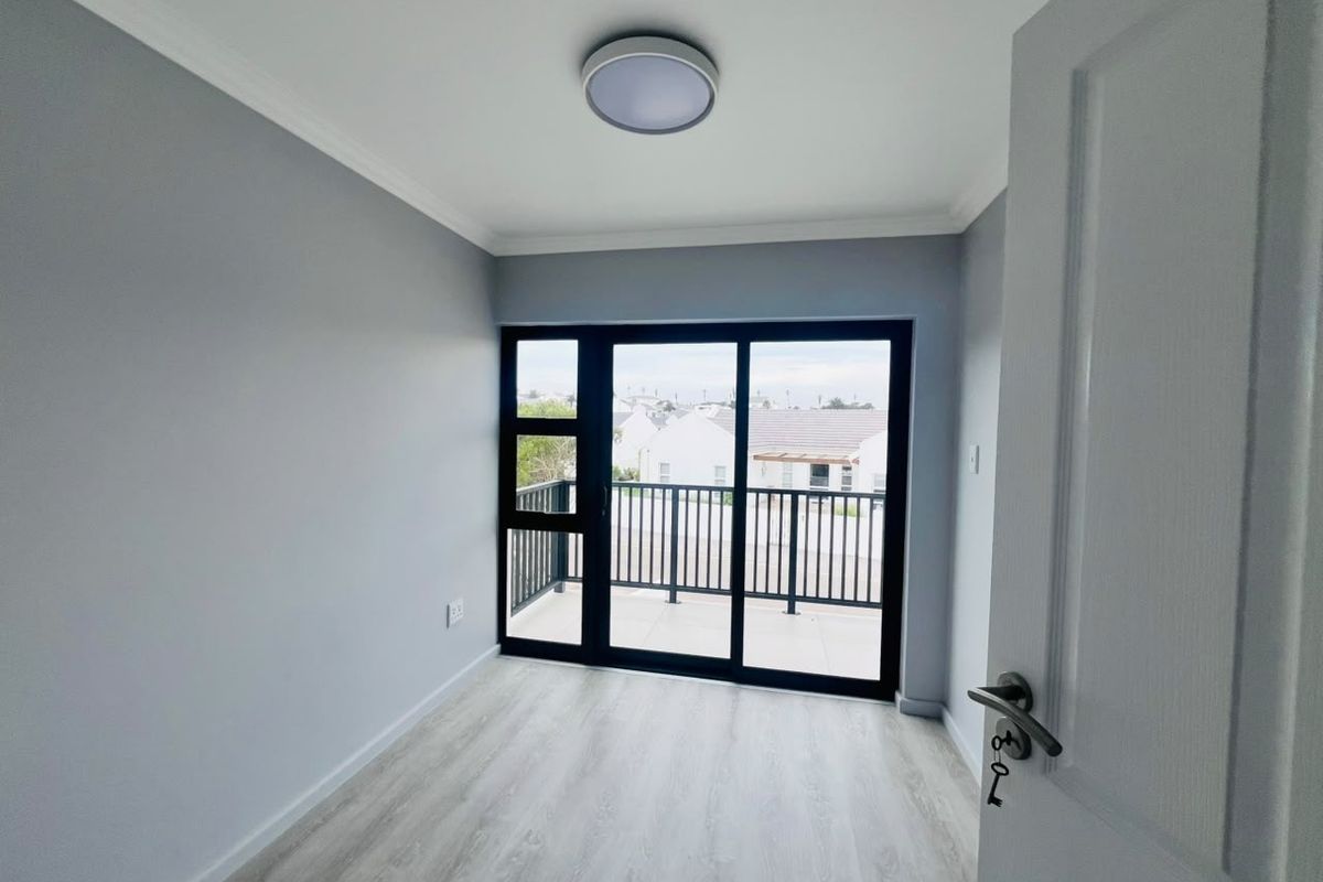 A bright, empty bedroom featuring modern laminate flooring, neutral grey walls, and a sliding glass door that opens onto a private balcony.