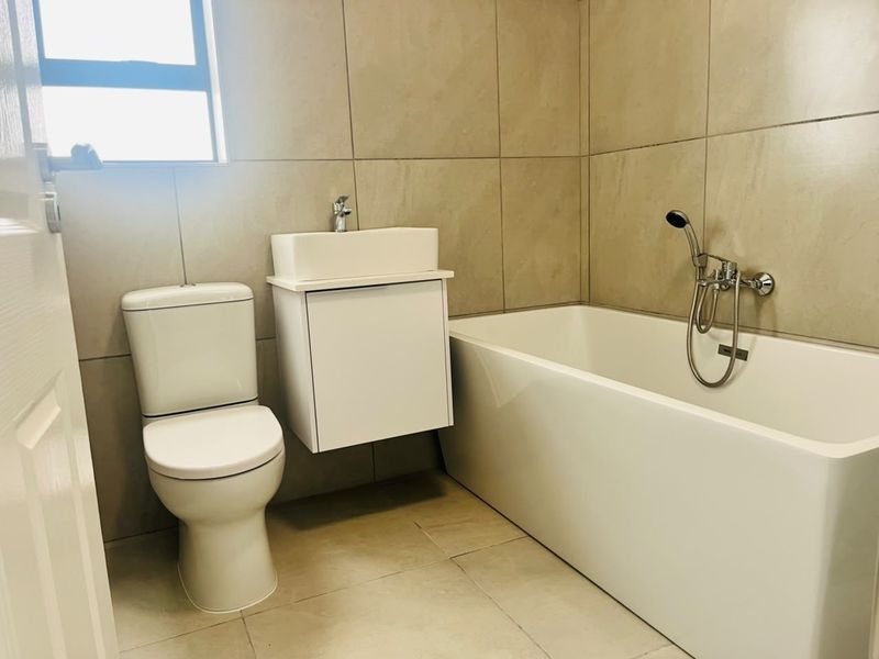 A modern, clean bathroom featuring neutral wall tiles, a wall-mounted vanity with a vessel sink, a standard toilet, and a bathtub with a shower mixer.