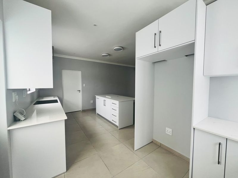 A modern, minimalist kitchen featuring white cabinetry, stone countertops, a central island, and neutral tiled flooring in an open-plan layout.