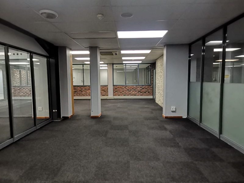 Spacious open-plan office area featuring glass partitioning, exposed brick accents, carpeted flooring, and integrated ceiling lighting with central air conditioning.