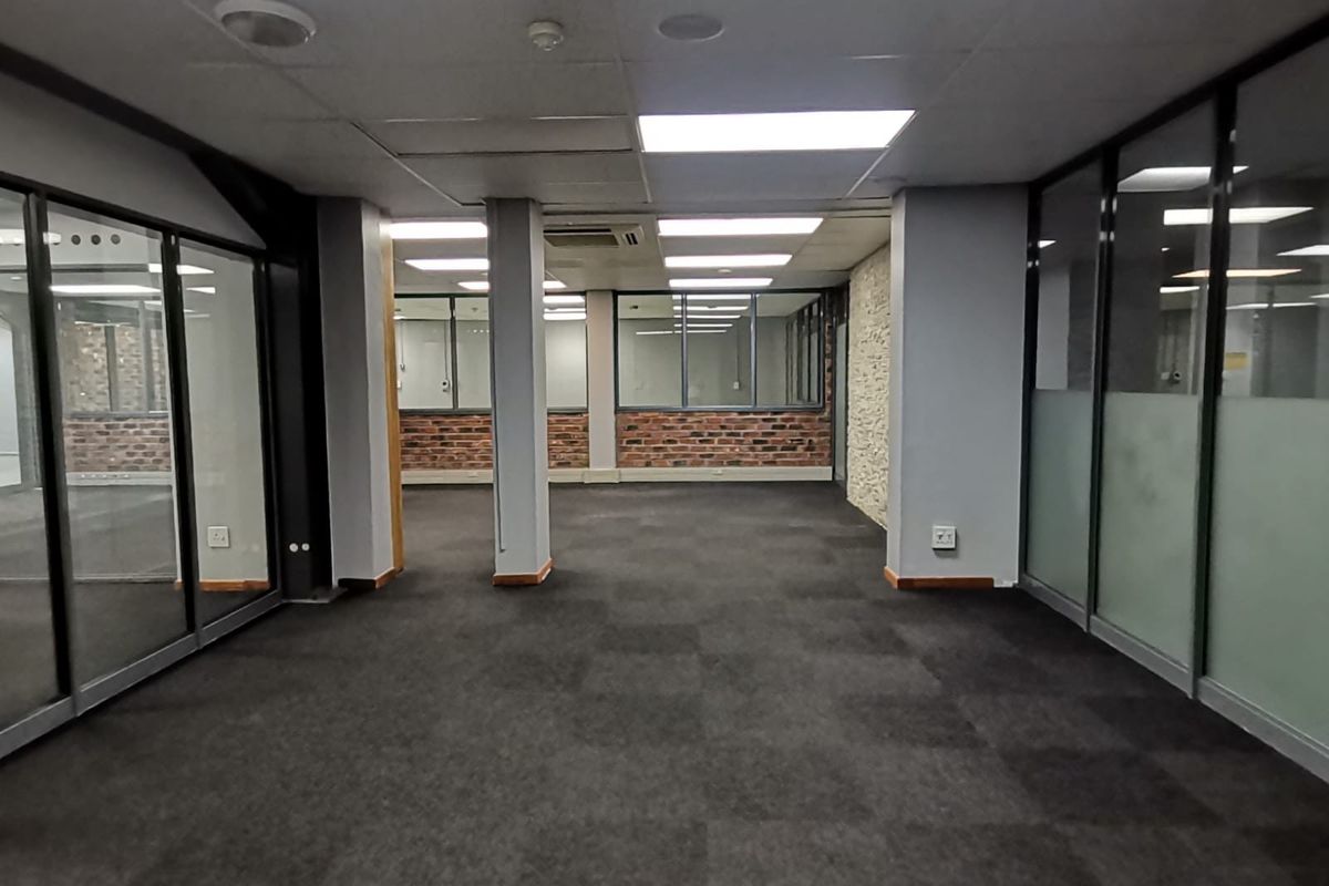 Spacious open-plan office area featuring glass partitioning, exposed brick accents, carpeted flooring, and integrated ceiling lighting with central air conditioning.