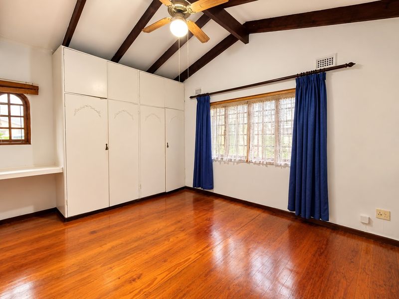 Spacious bedroom featuring polished timber flooring, exposed ceiling beams, built-in cupboards, and a built-in desk area with natural light.