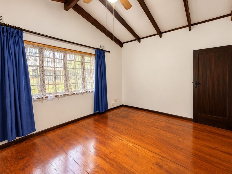 A bright, empty bedroom featuring polished timber flooring, exposed wooden ceiling beams, a ceiling fan, and a window with security bars.