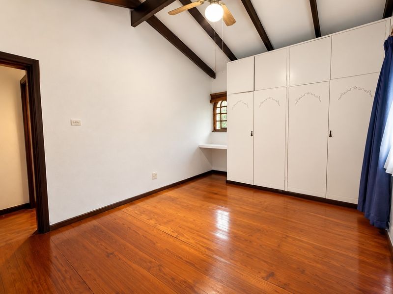 A spacious bedroom featuring polished timber flooring, exposed ceiling beams, a ceiling fan, and extensive built-in cupboard storage with an integrated desk area.