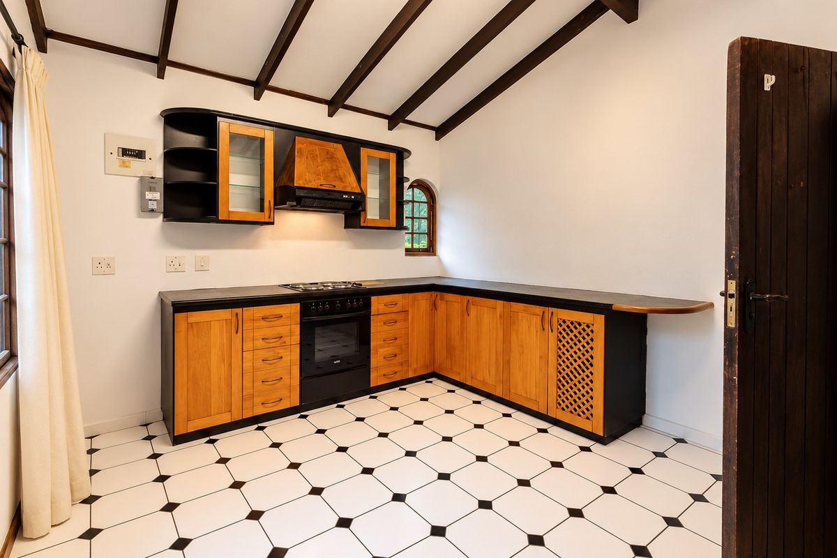 A well-maintained kitchen featuring wooden cabinetry, black countertops, built-in oven and hob, exposed timber ceiling beams, and classic black and white tiled flooring.
