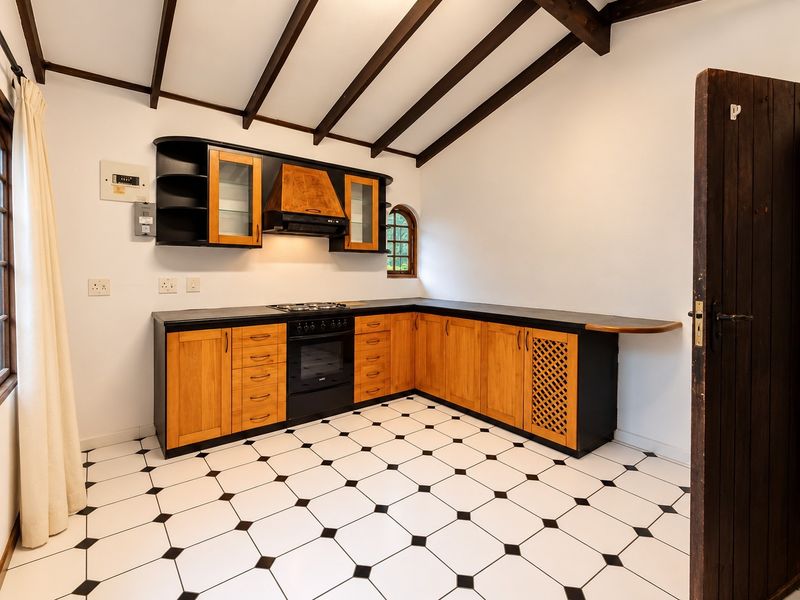A well-maintained kitchen featuring wooden cabinetry, black countertops, built-in oven and hob, exposed timber ceiling beams, and classic black and white tiled flooring.