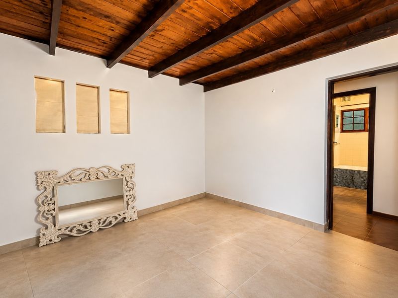 A bright, empty bedroom featuring exposed timber ceiling beams, neutral tiled flooring, and direct access to an adjoining bathroom.